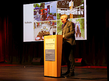 Norbert Seidl hinter dem Rednerpult auf der Bühne, im Hintergrund sind auf der Leinwand Fotos zu sehen.