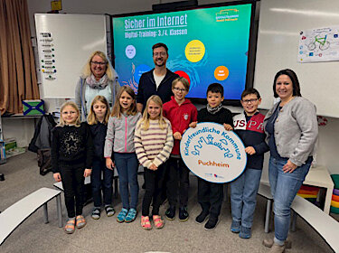 Gruppenbild mit Kindern, Rektorin, Trainer und der Vorsitzenden des Elternbeirats. Die Kinder halten ein großes Log der Kinderfreundlichen Kommune Puchheim.