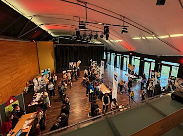 Blick von oben auf den Saal mit verschiedenen Infoständen und Besucher:innen Blick von oben auf den Saal mit verschiedenen Infoständen und Besucher:innen